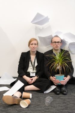 Two office workers in suits sat against a wall. They look confused. One is holding a plant. There is paper everywhere.