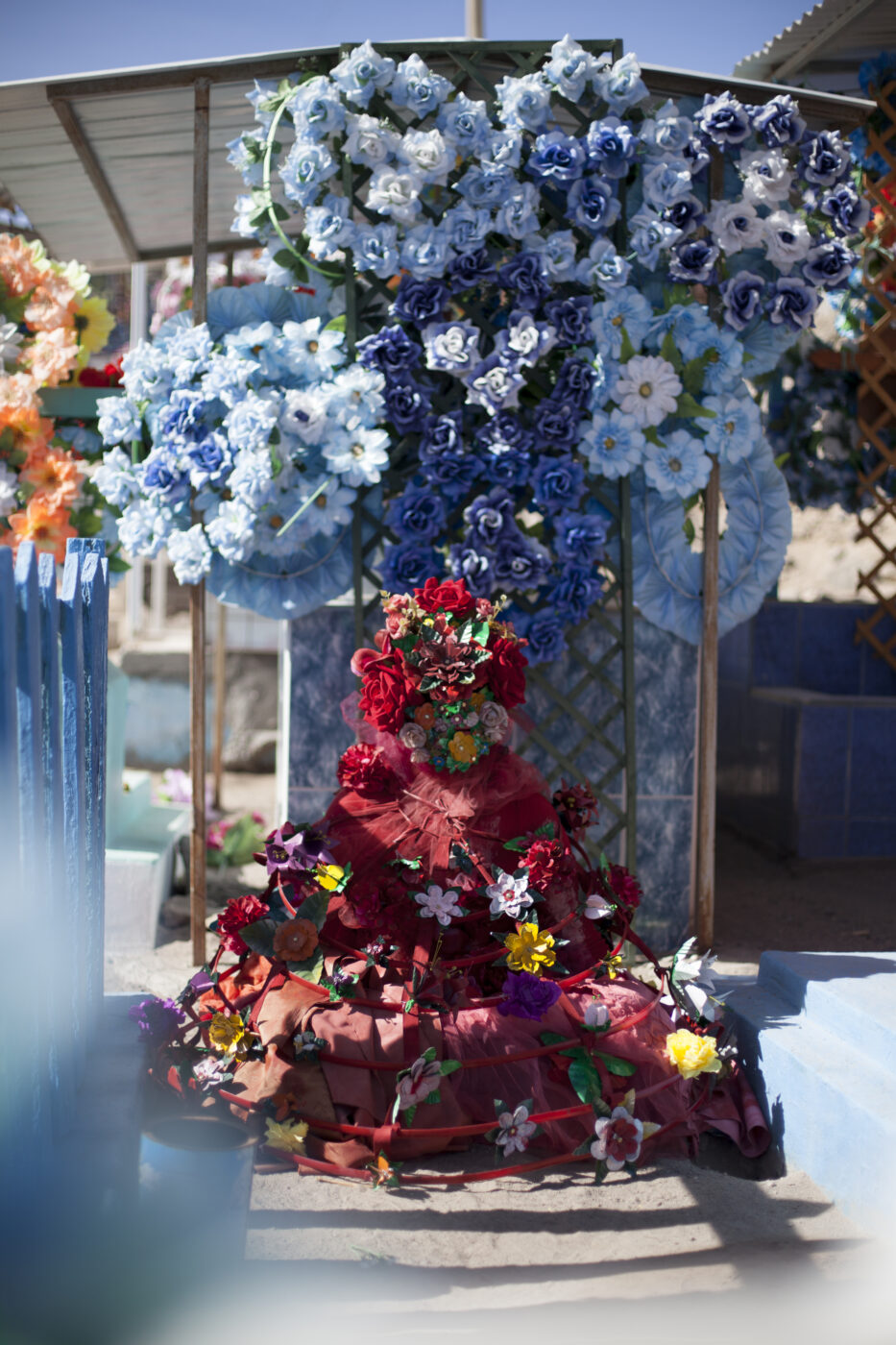 Alma y Muertos. Flower arrangement blues and yellows across the top and a pyramid of red and pink flowers rising from the front middle of the image.